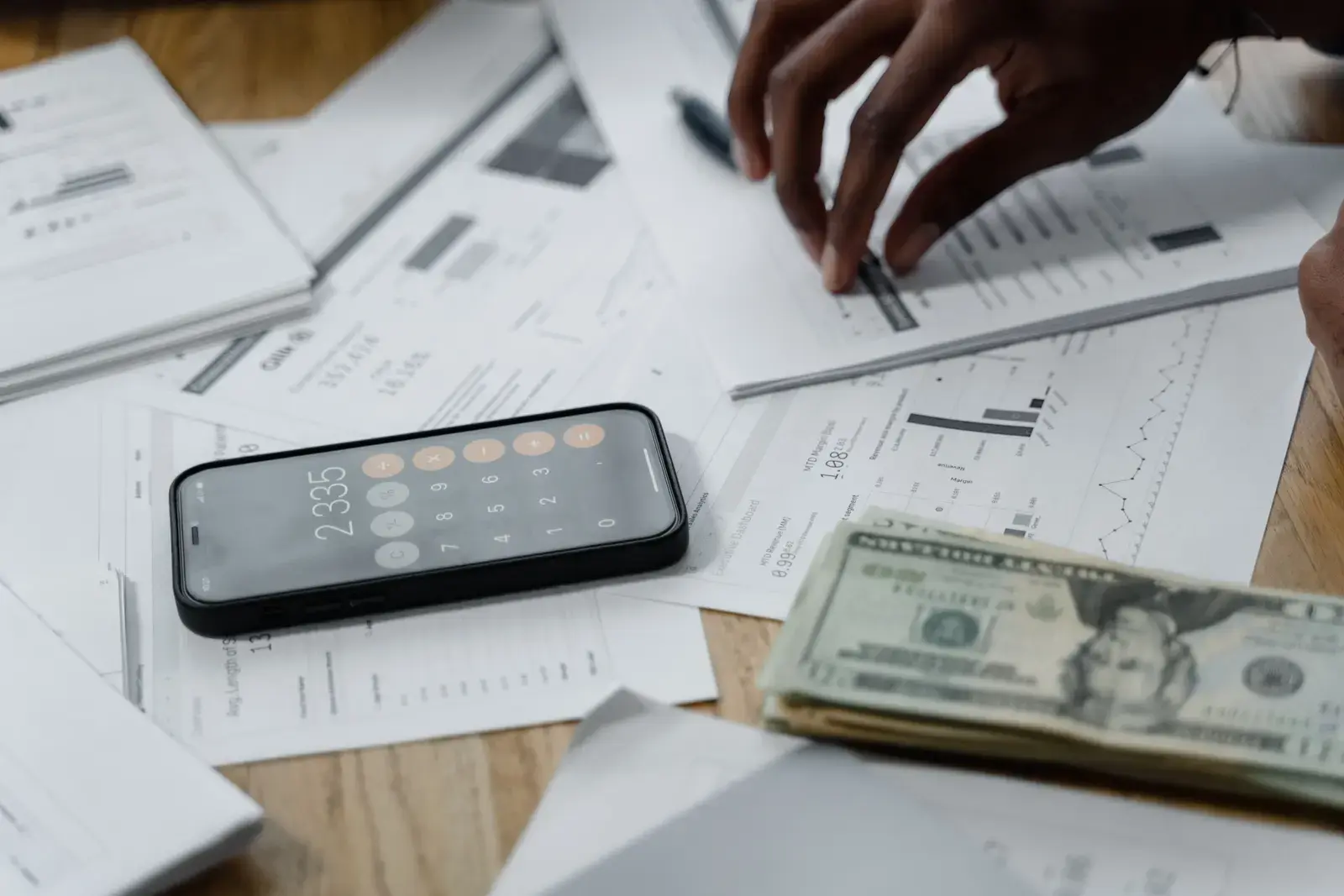 A hand sorting printed financial reports beside a phone calculator and banknotes