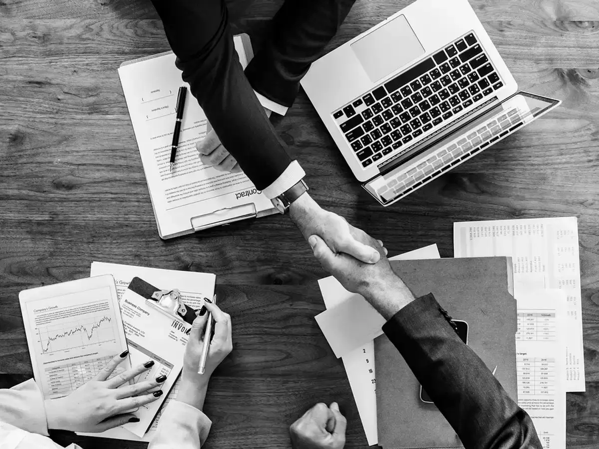 Two people shaking hands across a table covered with documents and a laptop